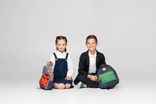 Cheerful schoolkids in uniform sitting with backpacks on grey — Stock Photo