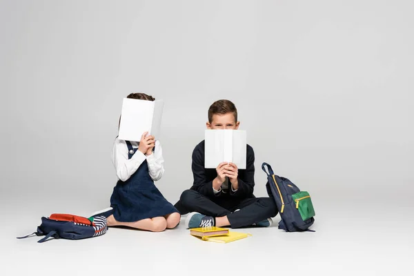 Schulkinder in Uniform sitzen da und bedecken Gesichter mit Büchern neben Rucksäcken auf grau — Stockfoto