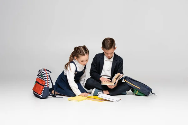 Schoolkids in uniform sitting and reading books near backpacks on grey — Stock Photo