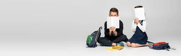 Schoolkids in uniform sitting and covering faces with books near backpacks on grey, banner — Stock Photo