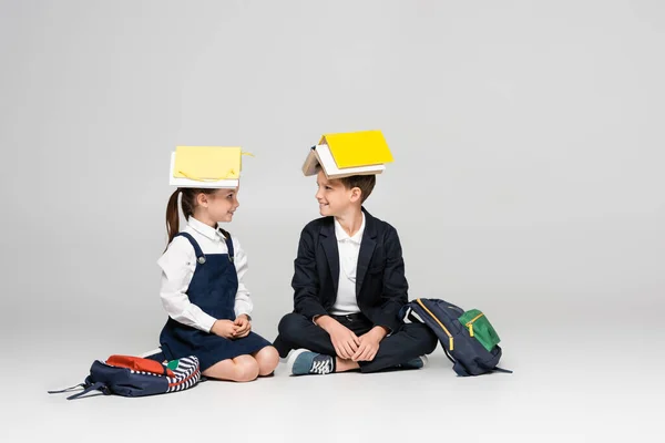 Happy schoolkids in uniform with books on heads sitting and looking at each other on grey — Stock Photo