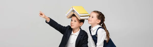 Школярка з книгами на голові вказує геть пальцем біля веселої дівчини ізольована на сірому, банер — стокове фото