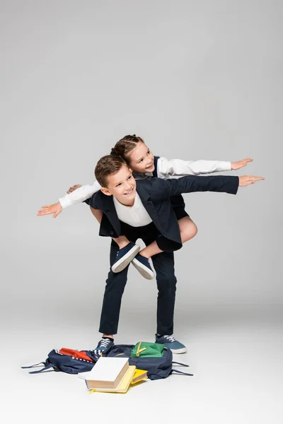 Happy schoolboy piggybacking friend with outstretched hands on grey — Stock Photo