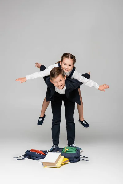 Cheerful schoolboy piggybacking girl with outstretched hands on grey — Stock Photo