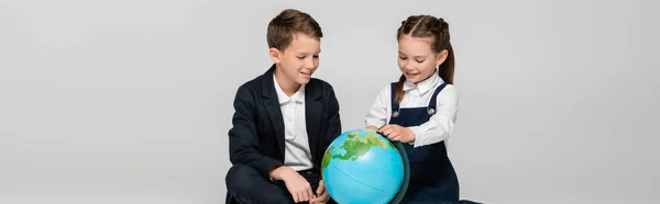 Happy schoolgirl and schoolboy looking at globe isolated on grey, banner — Stock Photo