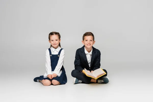 Positive schoolkids sitting with books and smiling on grey — Stock Photo