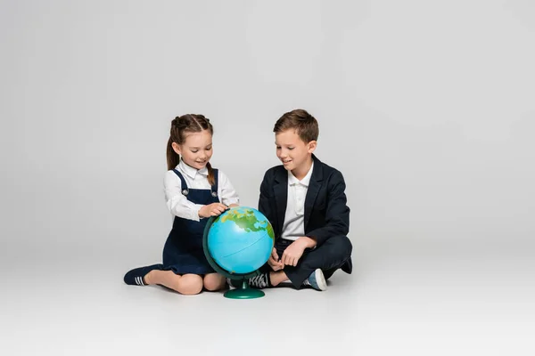Happy schoolgirl and schoolboy looking at globe while sitting on grey — Stock Photo