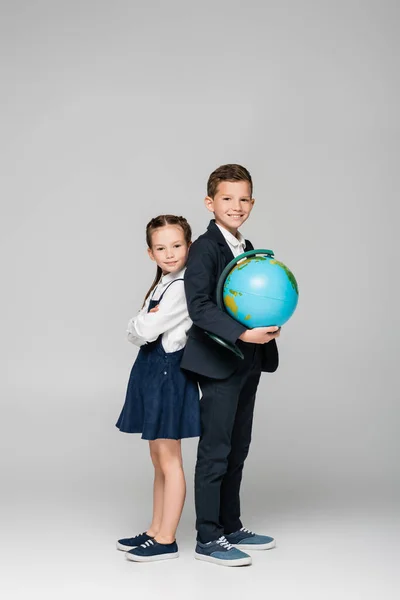 Щасливий школяр тримає глобус, стоячи з дівчиною в одязі на сірому — стокове фото