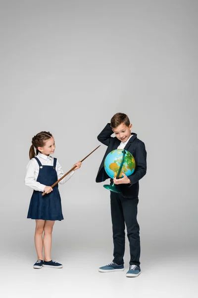 Улыбающаяся школьница держит палку рядом с запутавшимся мальчиком с глобусом на сером — стоковое фото