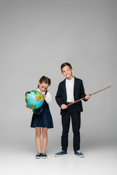 Cheerful schoolboy holding pointing stick near sad girl in dress with globe on grey — Stock Photo