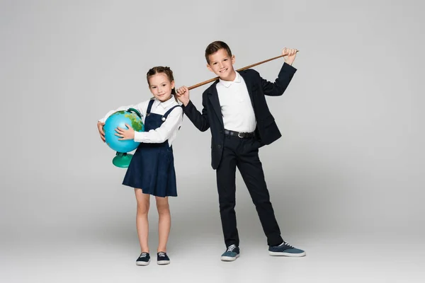 Feliz colegial sosteniendo apuntando palo cerca sonriente chica en vestido con globo en gris - foto de stock