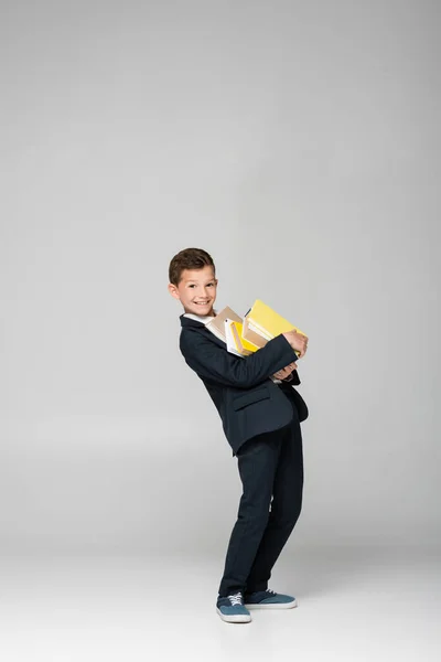 Feliz colegial en blazer y pantalones sosteniendo pila de libros en gris - foto de stock