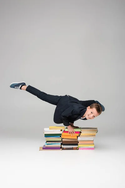 Happy schoolboy doing handstand on pile of books on grey — Stock Photo