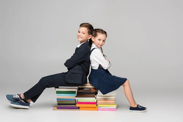Happy schoolkids sitting on pile of books on grey — Stock Photo