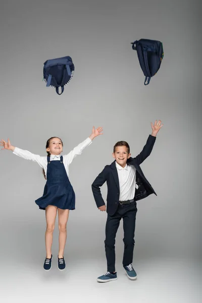 Glückliche Schulkinder werfen Rucksäcke in die Luft auf grau — Stockfoto