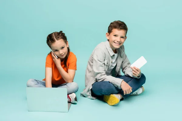 Girl talking on smartphone and using laptop near boy on blue — Stock Photo