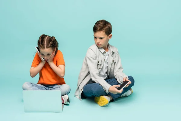 Shocked girl talking on smartphone and using laptop near sad boy on blue — Stock Photo