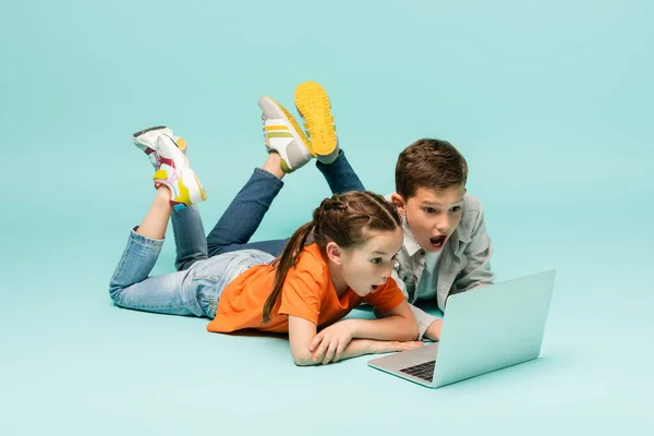 Schockierte Kinder sehen Film auf Laptop, während sie auf blauem Grund liegen — Stockfoto