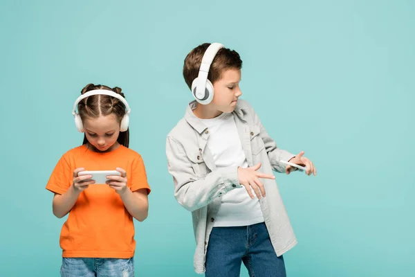 Girl using smartphone near boy in wireless headphones listening music while dancing isolated on blue — Stock Photo