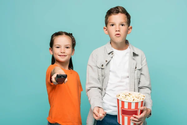 Girl holding remote controller near boy with popcorn bucket isolated on blue — Stock Photo