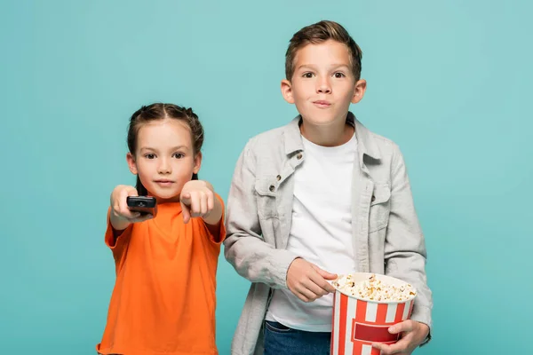 Дівчина тримає пульт дистанційного керування і вказує пальцем біля хлопчика з попкорн відром ізольовано на синьому — стокове фото