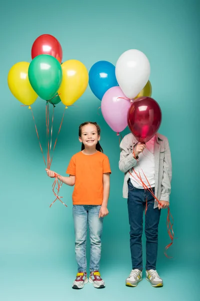 Menina feliz segurando balões coloridos perto do menino obscurecendo o rosto no azul — Fotografia de Stock