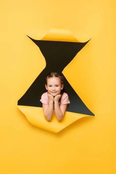 Happy girl looking at camera through hole on yellow background — Stock Photo