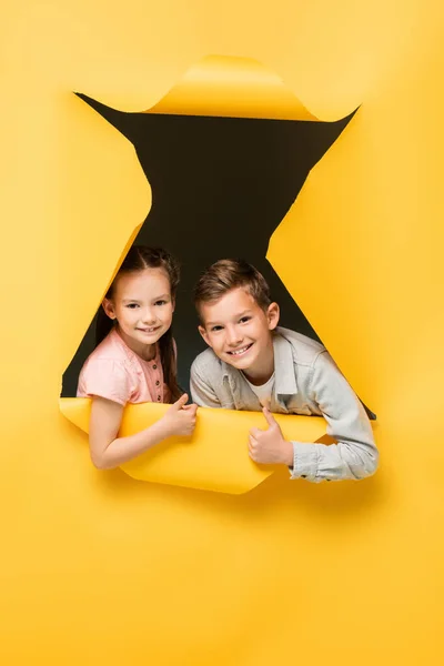 Happy kids showing thumbs up and looking at camera through hole on yellow background — Stock Photo