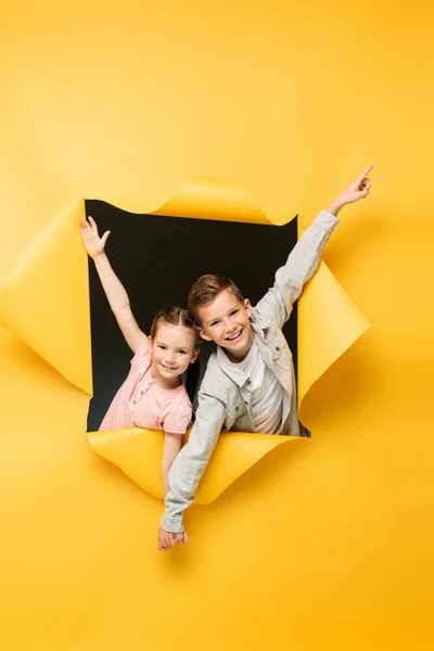 Glückliche Kinder, die Händchen halten und durch ein Loch auf gelbem Hintergrund lächeln — Stockfoto