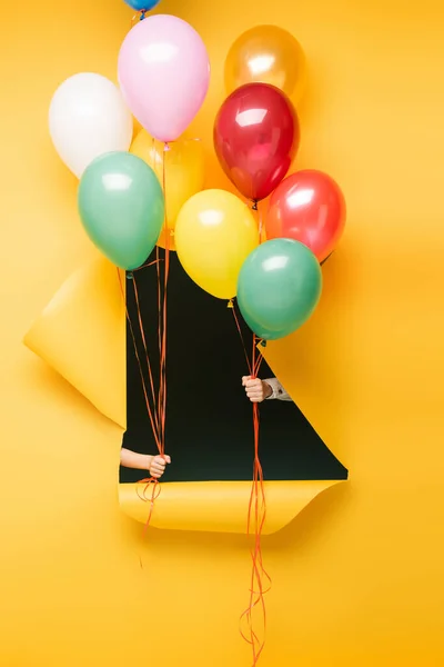 Abgeschnittene Ansicht von Kindern, die bunte Luftballons durch ein Loch auf gelbem Hintergrund halten — Stockfoto