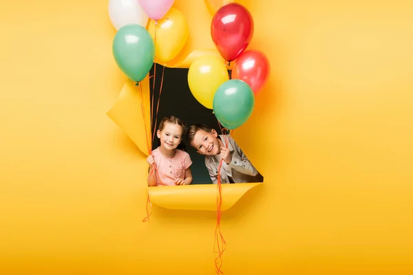 Cheerful kids holding colorful balloons through hole on yellow background — Stock Photo