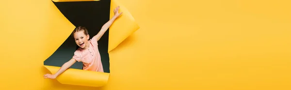 Збуджена дівчина з розтягнутими руками, дивлячись на камеру через рваний отвір на жовтому фоні, банер — стокове фото