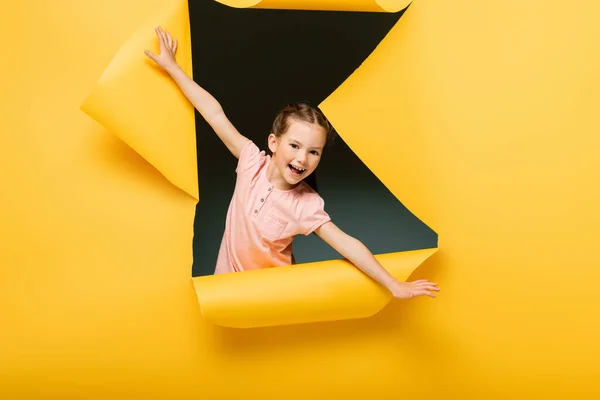 Excited kid with outstretched hands looking at camera through ripped hole on yellow background — Stock Photo