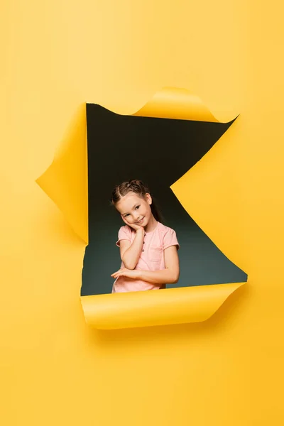 Pleased kid looking at camera while smiling through ripped hole on yellow background — Stock Photo