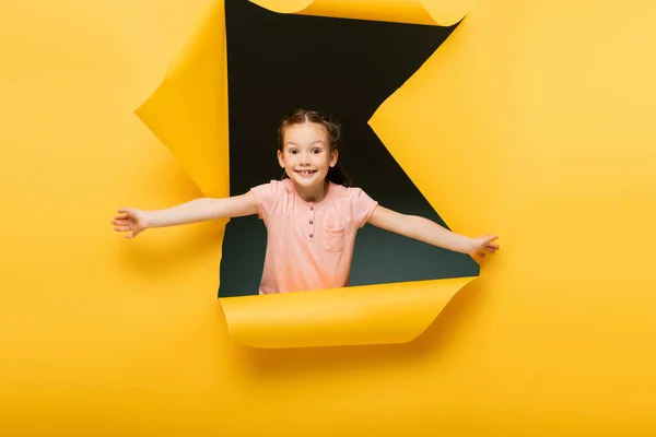 Niño alegre con las manos extendidas mirando a la cámara a través de un agujero rasgado en el fondo amarillo - foto de stock