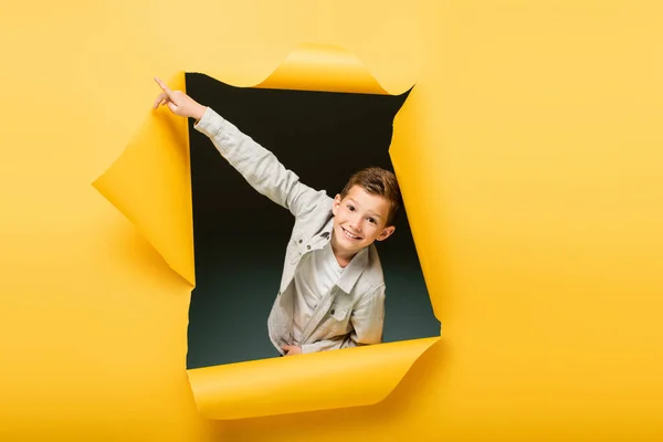 Happy boy pointing with finger and looking at camera through ripped hole on yellow background — Stock Photo