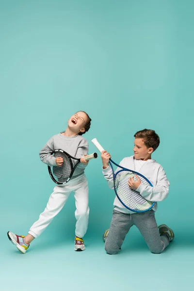 Enfants stupéfaits tenant des raquettes de tennis tout en chantant et en faisant semblant de jouer sur le bleu — Photo de stock