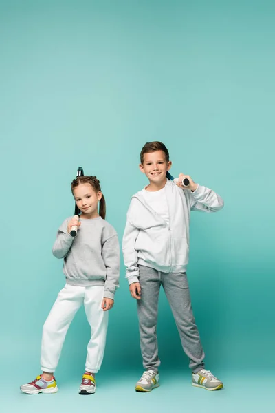 Sportive children in sportswear standing with tennis rackets and smiling on blue — Stock Photo