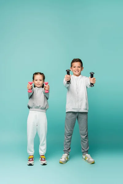 Niños felices en ropa deportiva y zapatillas de deporte trabajando con pesas en azul - foto de stock