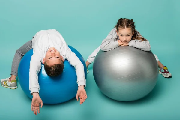 Счастливый мальчик в костюме лежа на фитнес-мяче рядом с девушкой на голубом — стоковое фото