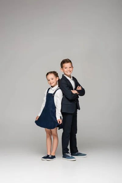 Feliz colegial de pie con brazos cruzados cerca de chica en vestido en gris - foto de stock