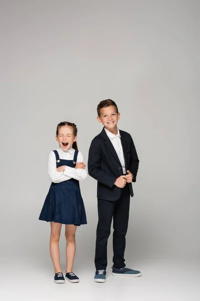 Colegiala en vestido bostezando cerca de niño en uniforme en gris - foto de stock