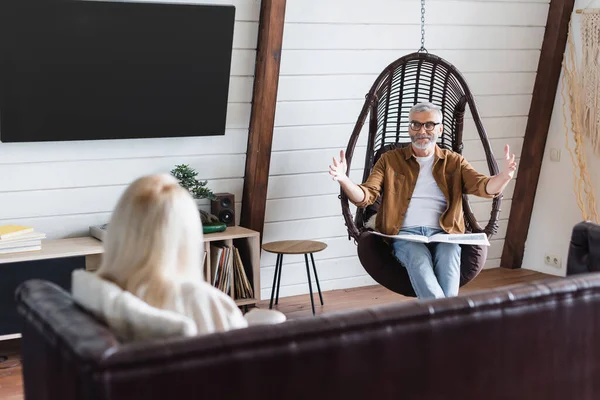 Fröhlicher Senior mit Zeitungspapier zeigt mit Händen neben verschwommener Frau auf Couch — Stockfoto