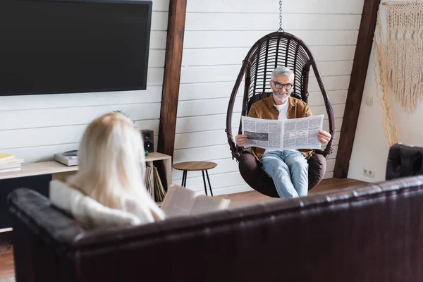 Senior liest Nachrichten im Hängesessel neben verschwommener Frau mit Buch im Wohnzimmer — Stockfoto