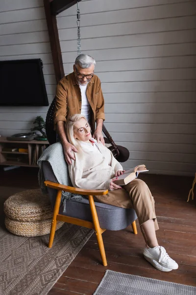 Старший мужчина обнимает жену с книгой в кресле — стоковое фото
