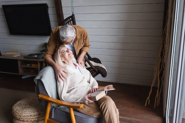 Anciano abrazando sonriente esposa con libro en sillón en casa - foto de stock