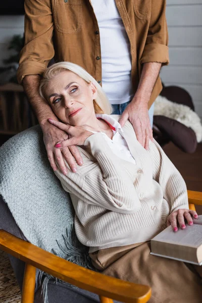 Seniorin mit verschwommenem Buch berührt Hand ihres Mannes zu Hause — Stockfoto