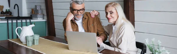 Couple âgé souriant utilisant un ordinateur portable sur la table de cuisine, bannière — Photo de stock