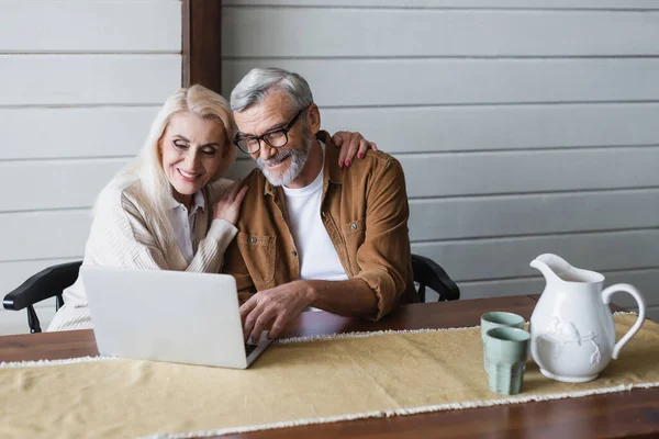 Elderly woman embracing smiling husband near laptop on kitchen table — Stock Photo