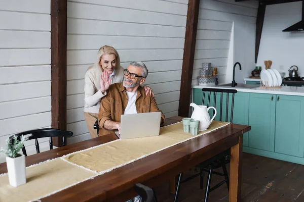 Smiling senior woman waving hand at laptop during video call near husband in kitchen — Stock Photo